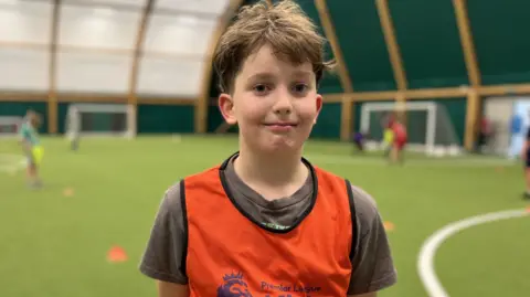 Noah (11) with short brown hair standing on the edge of an indoor football pitch wearing a bright orange Premier League Kicks bib.